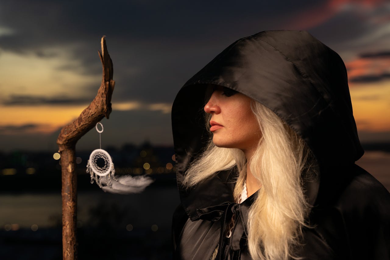 Menu A woman in a black hood holds a dreamcatcher at sunset, exuding mystery.