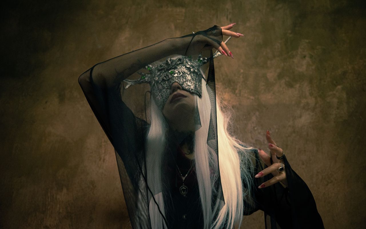 Menu A person with long white hair in an ornate mask poses dramatically against a textured backdrop.